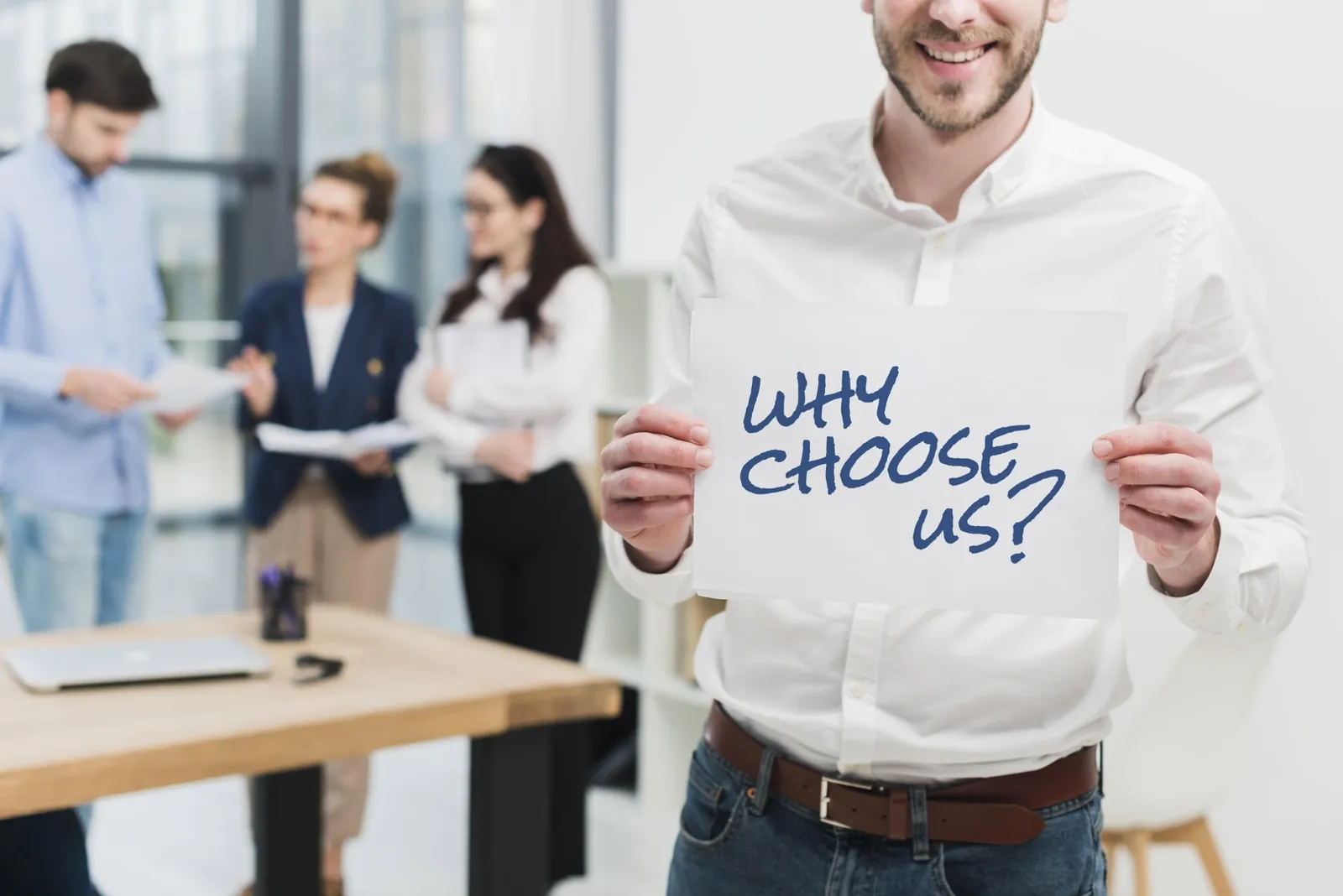 Un homme souriant tient une feuille où est écrit « Why choose us? » ; une équipe discute derrière lui.