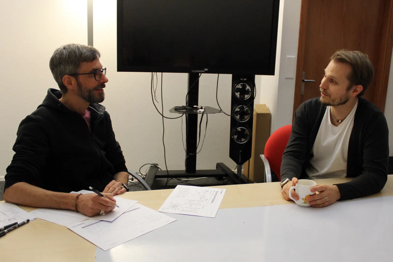 Deux hommes discutent à une table de réunion, entourés de feuilles de questionnaire.